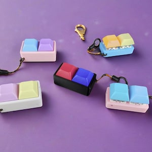May include: Five colorful keychains with different colored keycaps. Each keychain has a small metal ring for attaching to keys or bags. The keycaps are made of silicone and have a variety of colors, including pink, blue, yellow, red, and black.