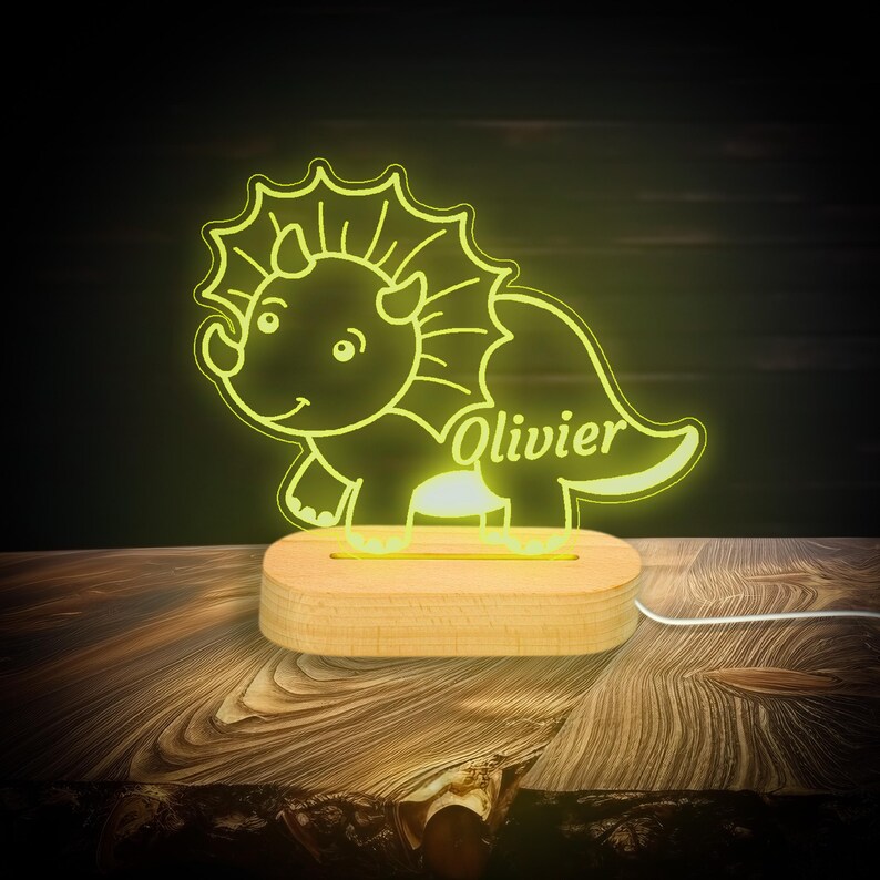 May include: A yellow night light shaped like a cartoon triceratops with the name "Olivier" written on the side. The light is on a wooden base.