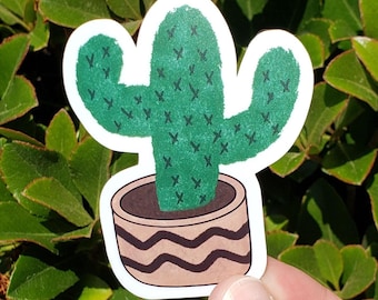 May include: A green cactus sticker in a brown pot with a wavy design. The cactus has a detailed texture and is set against a background of green leaves. The sticker is held by a hand.