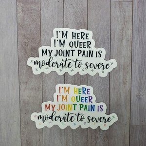 May include: Two white stickers with rainbow text that reads "I'm here I'm queer my joint pain is moderate to severe" with a few small stars around the text.