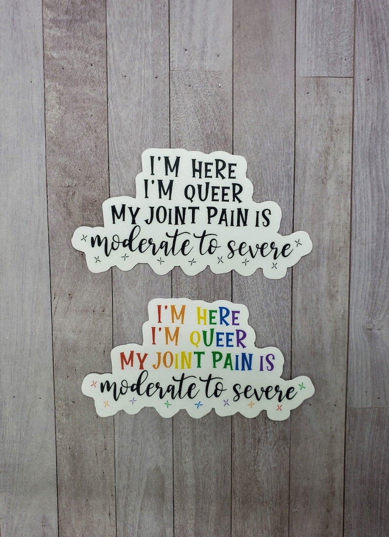 May include: Two white stickers with rainbow text that reads "I'm here I'm queer my joint pain is moderate to severe" with a few small stars around the text.