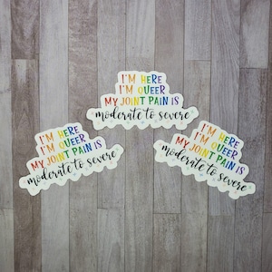 May include: Three rainbow-colored stickers with the text "I'm here, I'm queer, my joint pain is moderate to severe" on a wood background.
