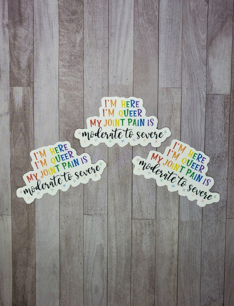 May include: Three rainbow-colored stickers with the text "I'm here, I'm queer, my joint pain is moderate to severe" on a wood background.