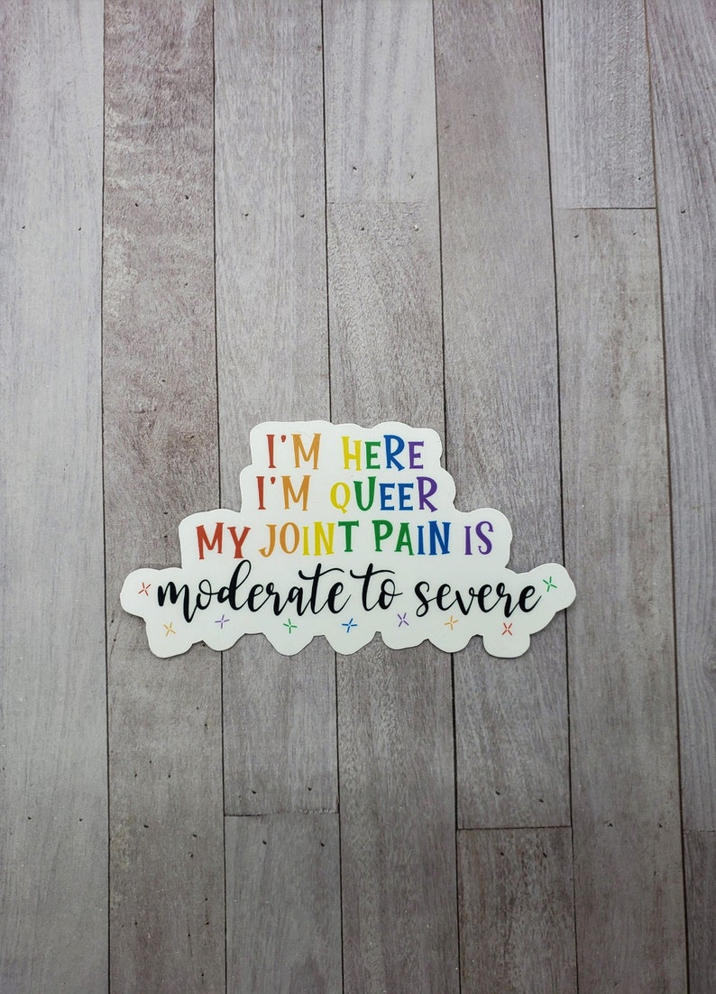 May include: A sticker with the text "I'm here, I'm queer, my joint pain is moderate to severe" in rainbow colors. The sticker is on a light wood background.