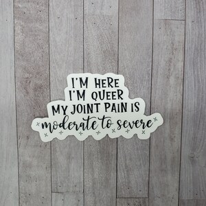 May include: White sticker with black text that reads "I'm here, I'm queer, my joint pain is *moderate to severe*" with stars around the text.
