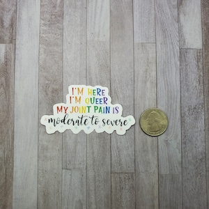 May include: A rainbow sticker with the text "I'm here I'm queer My joint pain is moderate to severe" on a wood background.