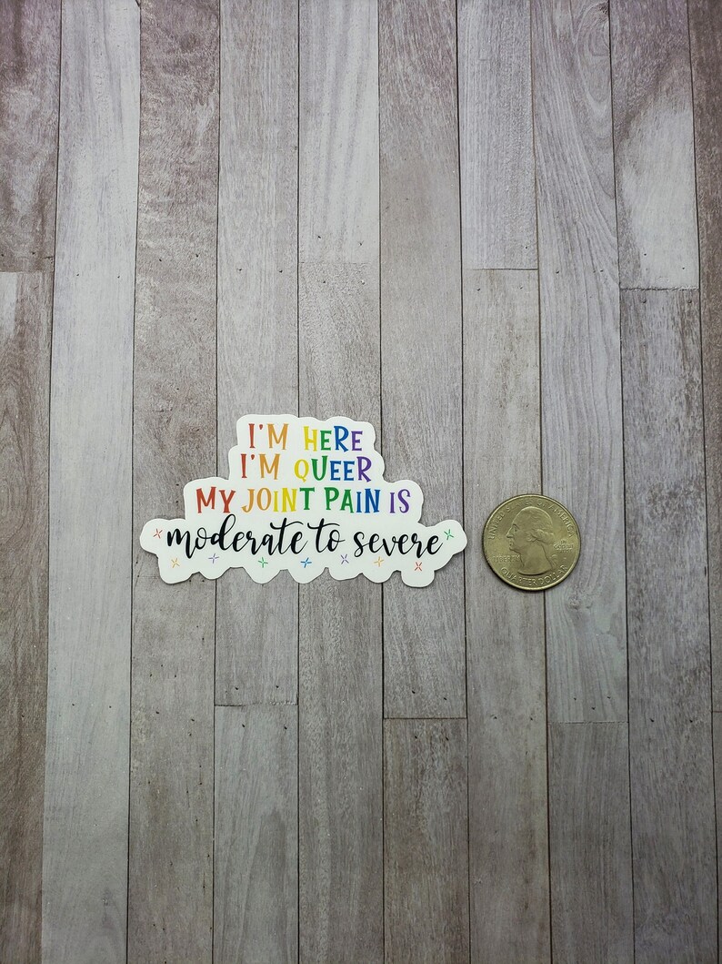 May include: A rainbow sticker with the text "I'm here I'm queer My joint pain is moderate to severe" on a wood background.