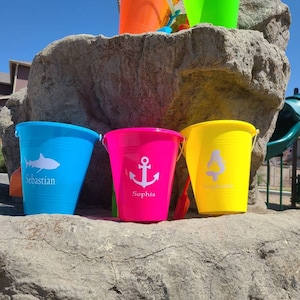 May include: A collection of colorful plastic beach buckets in orange, green, blue, pink, and yellow. Each bucket has a white graphic and a name: a shark with "Sebastian", an anchor with "Sophia", and a mermaid with "Stephanie".