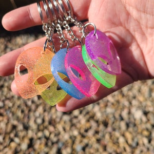 May include: A set of six colorful alien-shaped keychains with glitter embedded in the resin. The keychains are attached to silver key rings.