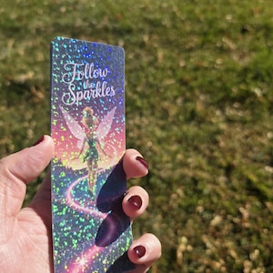May include: A rectangular bookmark with a holographic finish. The bookmark features a fairy illustration with the text "Follow the Sparkles." The fairy is depicted with wings and a flowing trail. The background has a gradient of colors.
