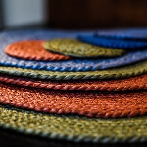 May include: A stack of round woven placemats in shades of green, blue, and orange. The placemats are made of a natural fiber and have a rope-like edge.