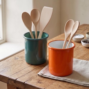 May include: Two enamel kitchen utensil holders, one teal and one orange, each filled with wooden spoons and a spatula. The holders sit on a wooden surface with small bowls of salt and pepper in the background. A linen napkin is under the orange holder.