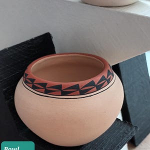 May include: A small, round, terracotta-coloured bowl with a decorative rim featuring a geometric pattern in black and red. The bowl is 9cm high and 12cm in diameter. Another bowl is visible in the background.