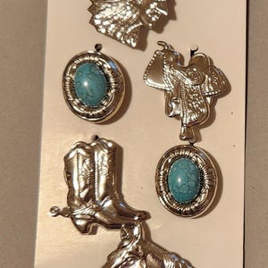 May include: A collection of silver-toned button covers with Western and Native American themes. Includes a headdress, a saddle, a cowboy boot, a horse head, and two round covers with turquoise-colored centers. The text "Button Covers" is at the top.