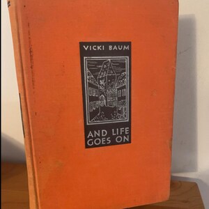 May include: An orange hardcover book titled "AND LIFE GOES ON" by Vicki Baum. The cover features a black and white illustration of a building. The book is displayed on a wooden surface.