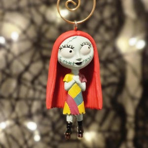 May include: A Sally ornament from The Nightmare Before Christmas. The figure has pale skin, red hair, and a patchwork dress in yellow, pink, and blue. The ornament is suspended by a gold wire. The figure's stitched features are visible.