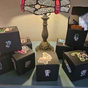 May include: Several black cube-shaped boxes with colorful character stickers are arranged on a table. One box is open, revealing a small notebook and figurines. A decorative lamp with a stained glass shade is in the background.
