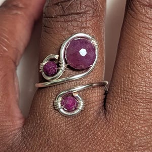 May include: A silver-toned ring with three faceted, deep pink gemstones. The ring features a swirling design with the gemstones set in wire-wrapped settings. The ring is on a finger.