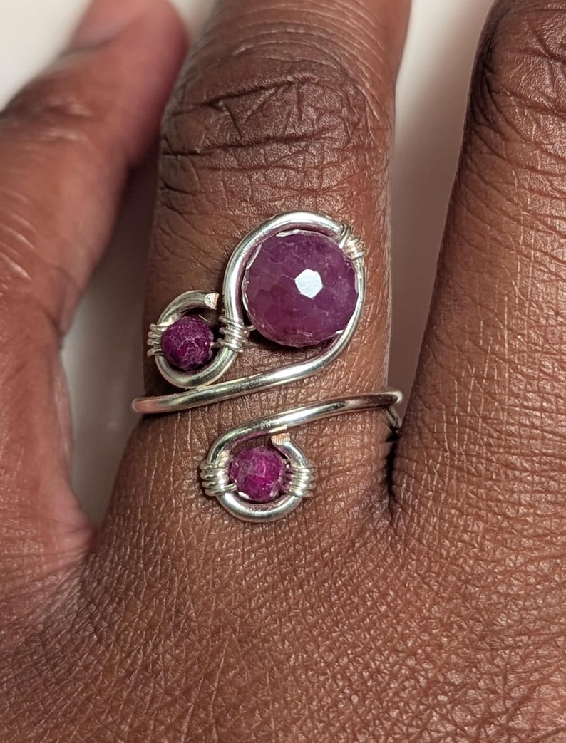 Op de afbeelding: Een zilverkleurige ring met drie gefacetteerde, dieproze edelstenen. De ring heeft een wervelend ontwerp met de edelstenen in draadomwikkelde zettingen. De ring zit om een vinger.