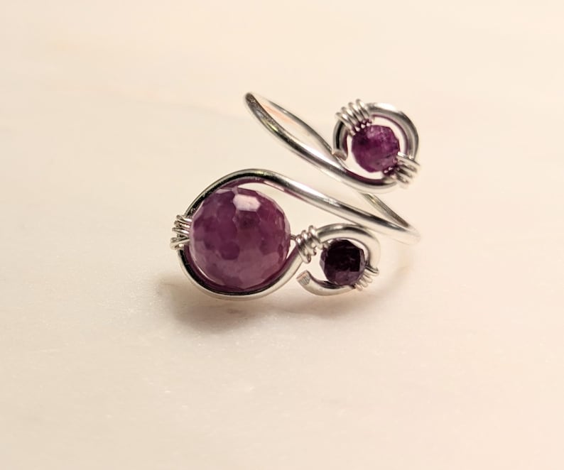 Op de afbeelding: Een zilverkleurige ring met drie gefacetteerde, dieppaarse edelstenen. Het ontwerp van de ring heeft een wervelende, open stijl, waarbij de edelstenen zijn vastgezet met draadwikkeling. De ring is geplaatst tegen een lichte achtergrond.
