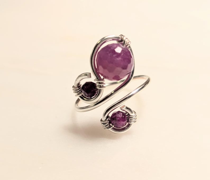 Op de afbeelding: Een zilverkleurige, draadgewikkelde ring met drie gefacetteerde, dieppaarse edelstenen. De ring heeft een wervelend ontwerp met een grote centrale steen en twee kleinere stenen aan de zijkanten.