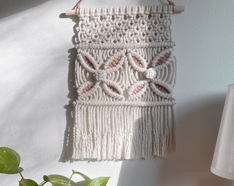 May include: A white macrame wall hanging with pink accents. The hanging is made with a knotting technique and has a fringe at the bottom. It is hanging on a white wall with a plant and a book in the foreground.