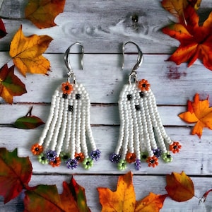 May include: A pair of white beaded ghost earrings with orange, green, and purple flower accents. The earrings are hanging from silver hooks.