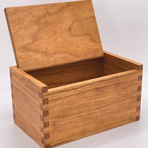 以下が含まれることがあります： 明るい茶色の木でできており、自然な木目がある、蝶番付きの蓋が付いた木製の箱。箱はシンプルで長方形の形をしており、鳩尾継ぎの構造が特徴です。