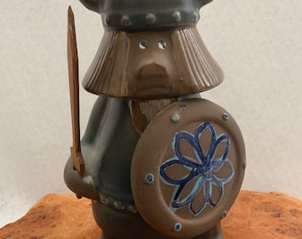 May include: A ceramic figurine of a Viking warrior wearing a horned helmet and holding a sword and shield. The shield has a blue and white flower design. The figurine is brown and gray with blue accents. The figurine is signed "Design Kurt Hlusson".