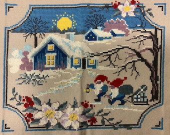 以下が含まれることがあります： 薄いベージュの布に施されたクロスステッチ刺繍。青い家、黄色い太陽、赤い帽子をかぶった2人のノームが描かれた冬の風景。1人のノームはランタンを持ち、もう1人は小さなそりを引いている。白い花と赤い実が周囲を飾っている。