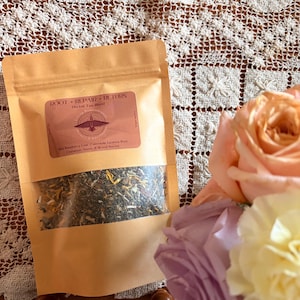 Könnte beinhalten: Ein beigefarbener Papierbeutel mit einem transparenten Fenster, gefüllt mit losem Kräutertee. Auf dem Etikett steht "ROOT • REPAIR • RETURN Herbal Tea Blend". Zu den Zutaten gehören rote Himbeerblätter, Ringelblume und Süßholzwurzel. Ein Strauß aus rosa und pfirsichfarbenen Rosen befindet sich im Hintergrund.