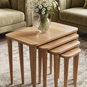 May include: A set of four light brown wooden nesting tables with rounded corners and tapered legs. The tables are arranged in a cascading formation, with a clear glass vase filled with white daisy flowers placed on the largest top table.