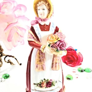 May include: A porcelain figurine of a woman wearing a red dress and a white apron, holding a basket of colorful roses. She is wearing a yellow hat and a plaid scarf.