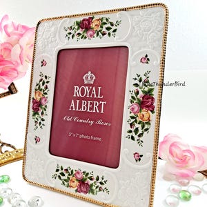 May include: White ceramic photo frame with gold trim and floral accents. The frame features the text "ROYAL ALBERT Old Country Roses" and is designed for a 5" x 7" photo. Decorative roses and leaves adorn the frame's corners.