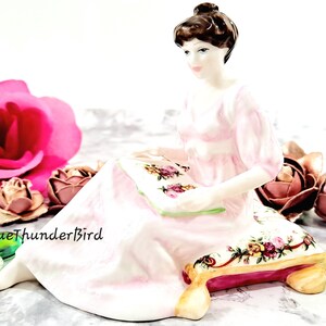 May include: A porcelain figurine of a woman in a pink dress sitting on a floral cushion, holding a book. The figurine is surrounded by pink and brown roses and green glass beads.