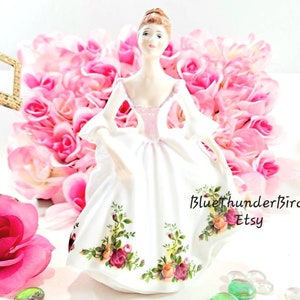 May include: A porcelain figurine of a woman wearing a white dress with pink and floral accents. The figurine is standing on a white surface with pink roses and green glass beads in the background. The text "BlueThunderBird Etsy" is visible in the bottom right corner of the image.