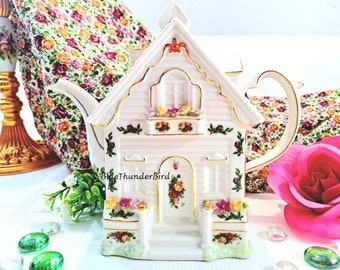 May include: White porcelain teapot shaped like a cottage with gold trim and floral details. The teapot has a spout and handle and is decorated with roses and greenery.