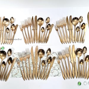 May include: A set of gold-toned silverware, including forks, knives, spoons, and serving utensils. The silverware is arranged in rows on a white surface.