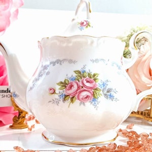 May include: A white teapot with a floral design featuring pink roses and blue forget-me-nots. The teapot has a gold trim and a scalloped edge. The teapot is sitting on a white surface with pink and peach colored rose petals.