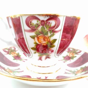 May include: A white teacup and saucer with a burgundy and white floral pattern. The pattern features roses and bows with gold trim.