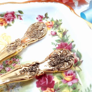 May include: Two gold-plated serving spoons with ornate, detailed designs. The spoons are on a white plate with a floral pattern.