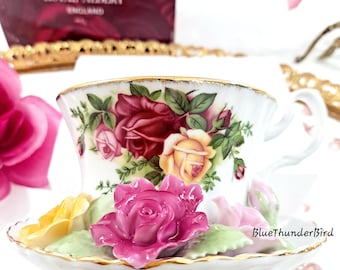 May include: A Royal Albert teacup and saucer set with a floral rose design in shades of red, pink, and yellow. The teacup and saucer have gold trim. A decorative rose is on the saucer. The box in the background says "Royal Albert England".