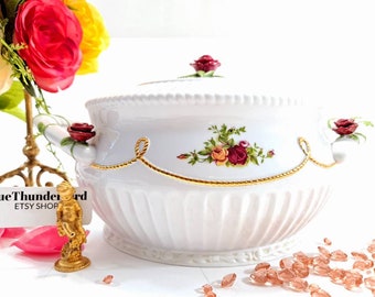 May include: White porcelain serving dish with a gold trim and floral design. The dish has a lid with a rosebud finial. The dish is on a white surface with pink glass beads.