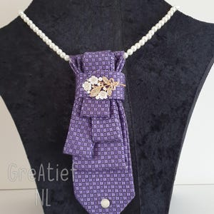 Puede incluir: Una corbata morada con un patrón cuadrado, adornada con un broche floral dorado y un collar de perlas. La corbata se exhibe en un maniquí negro, mostrando su diseño y detalles.