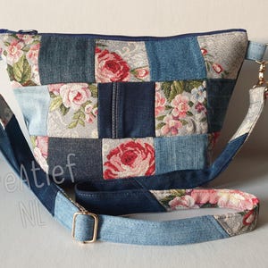 Op de afbeelding: Een patchwork crossbody tas met een bloemenontwerp en denim vierkanten. De tas heeft een mix van licht en donkerblauw denim, met een bloemenpatroon van roze rozen en groene bladeren. Het heeft een lange, verstelbare riem en goudkleurige hardware.