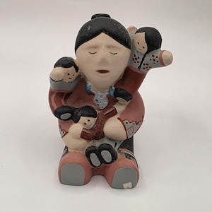 May include: A ceramic figurine depicting a larger figure with black hair and a red garment, holding three smaller figures. The larger figure wears a blue necklace. The smaller figures have black hair and are positioned on the shoulders and lap.