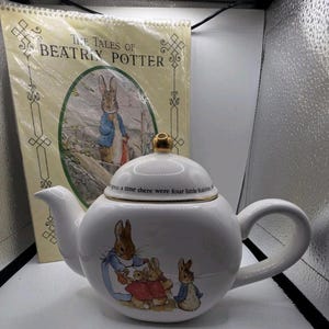 May include: A white ceramic teapot featuring illustrations of Peter Rabbit and family. The lid has a gold-colored knob and the text "Once upon a time there were four little Rabbits". Behind it is a book titled "The Tales of Beatrix Potter".