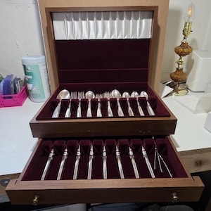 May include: A wooden flatware chest with a hinged lid and a pull-out drawer, both lined with deep burgundy velvet. The interior features silver-toned spoons, forks, and knives arranged in molded slots, with a white satin panel inside the lid.