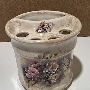 May include: A cream-colored ceramic toothbrush holder with a crackled glaze finish. The cylindrical container features a floral design with pink and muted brown roses on the front and a smaller floral cluster on the top rim, which has four holes.