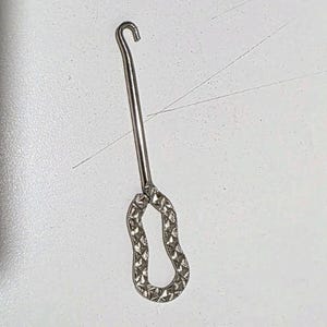 May include: A silver-toned metal button hook featuring a long, slender shaft that terminates in a curved hook at the top. The handle is shaped like an elongated, irregular oval and is finished with a textured, diamond-cut pattern.
